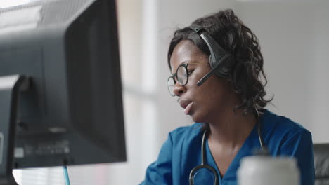 African-Female-medical-assistant-wears-white-coat-headset-video-calling-distant-patient-on-computer.-Doctor-talking-to-client-using-virtual-chat-computer-app.-Telemedicine-remote-healthcare